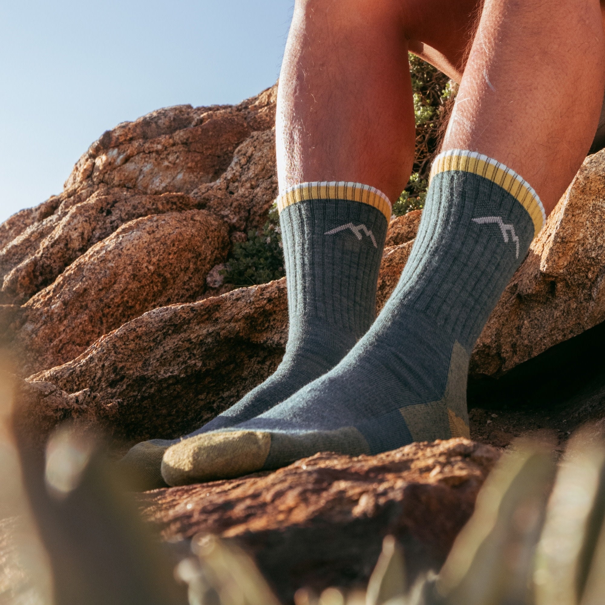 Crew hiking socks in teal with gold striped cuff and mountain logo, worn on rocky terrain