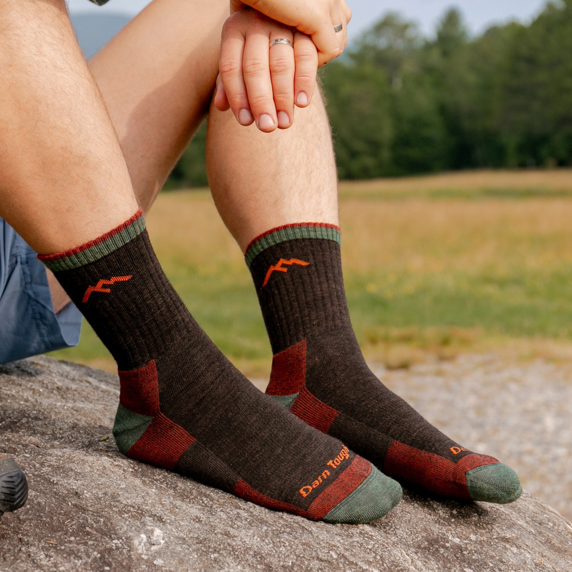 Crew-length hiking socks in dark brown with green cuff, burgundy heel and toe accents, worn on rock outdoors