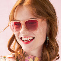 Pink framed sunglasses with red gradient lenses worn by smiling woman with freckles