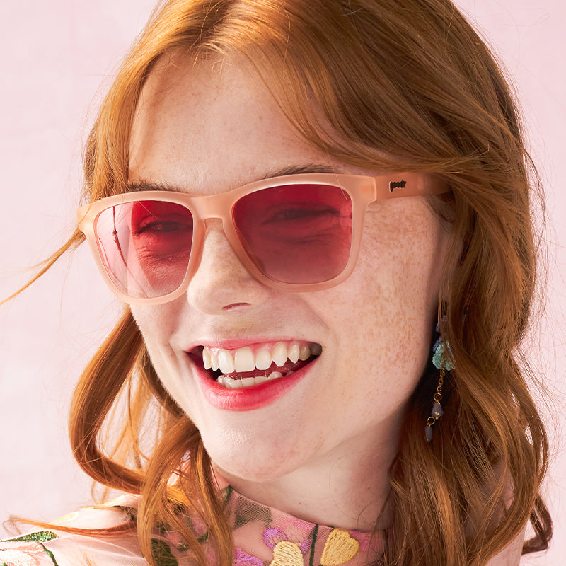 Pink framed sunglasses with red gradient lenses worn by smiling woman with freckles