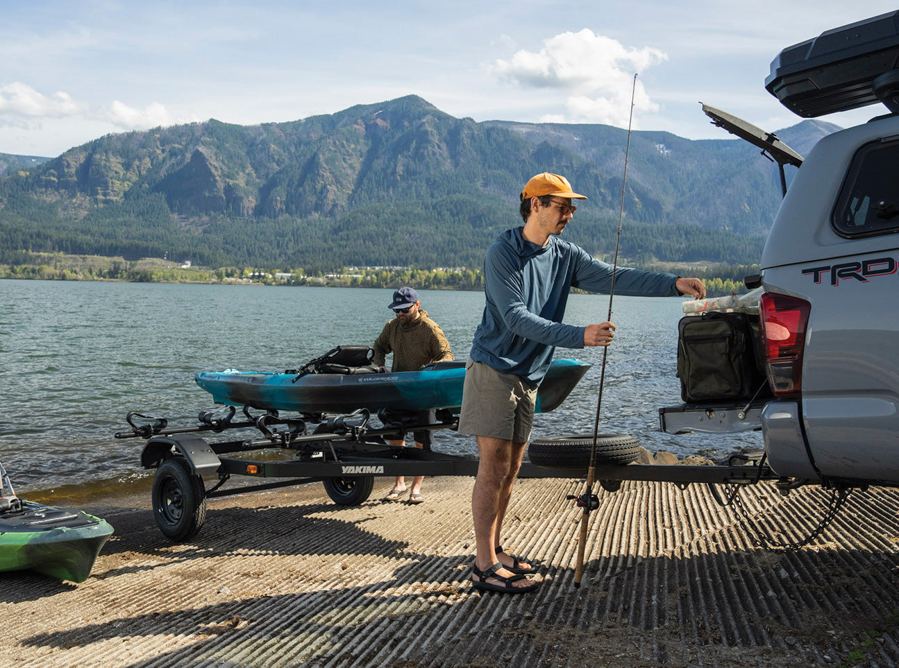 Black kayak trailer with multiple kayaks loaded, man preparing fishing gear beside pickup truck at lake