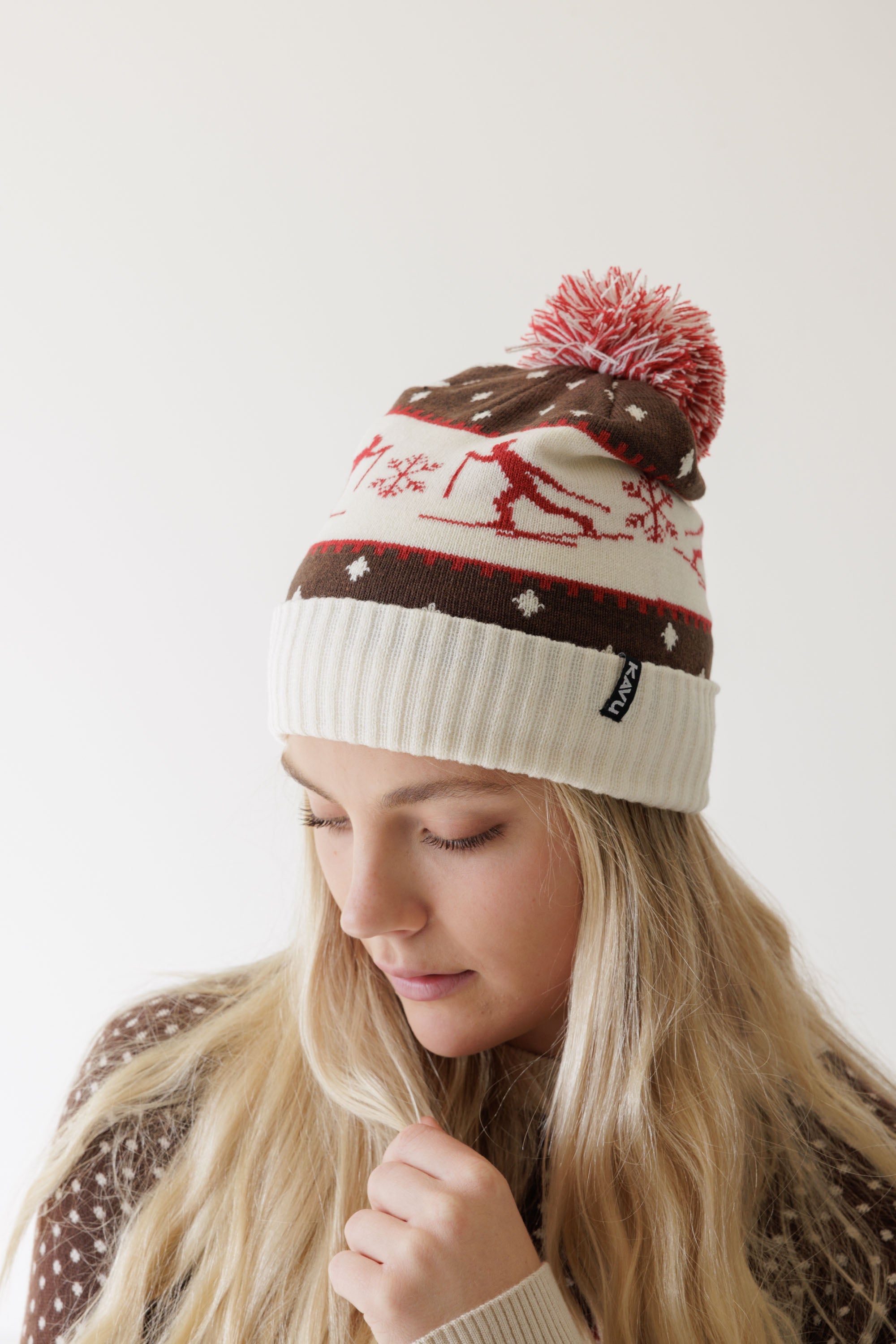 Knit winter beanie with nordic reindeer pattern, red and white pom-pom, worn by model looking down