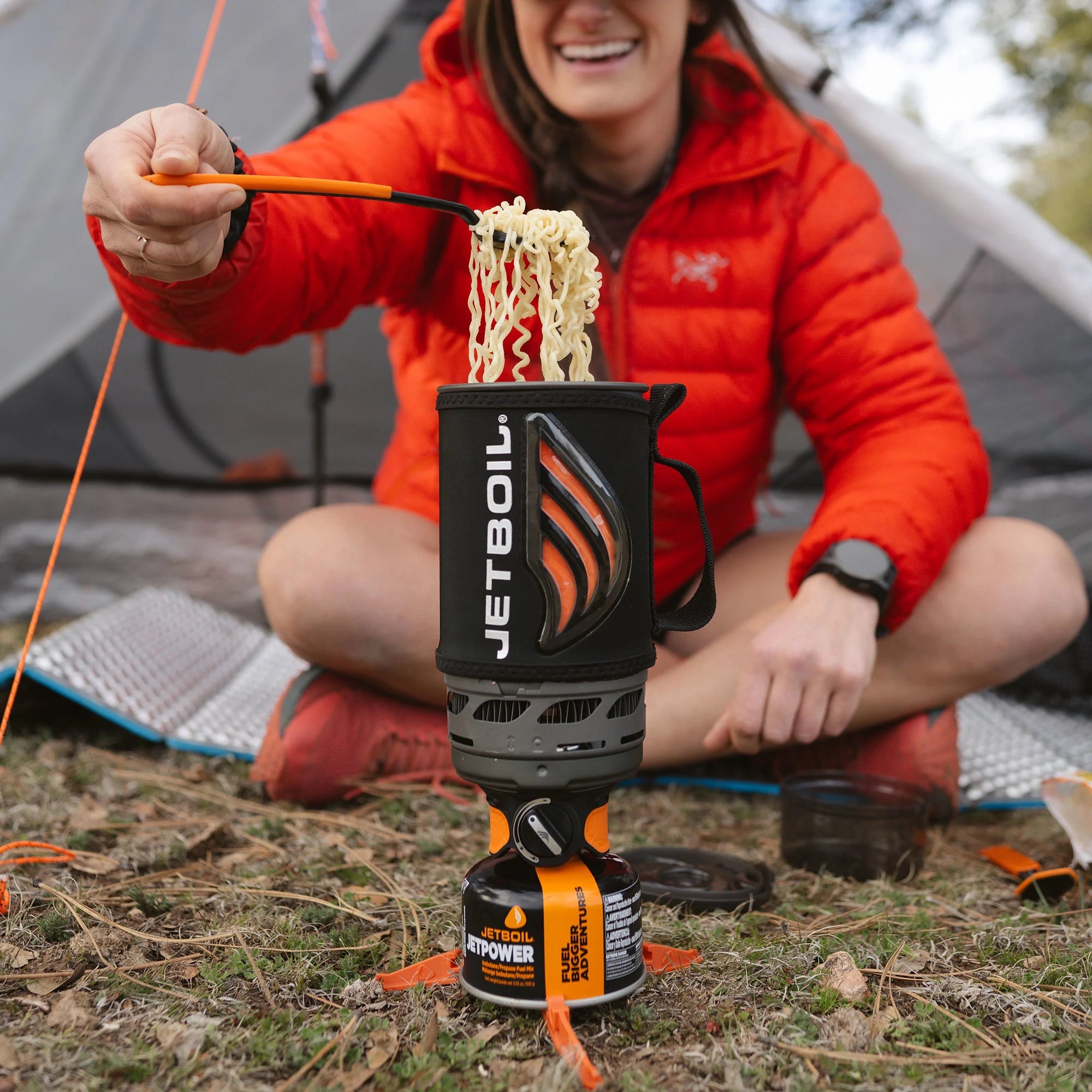Compact camping stove system with noodles being lifted by chopsticks, mounted on fuel canister outdoors