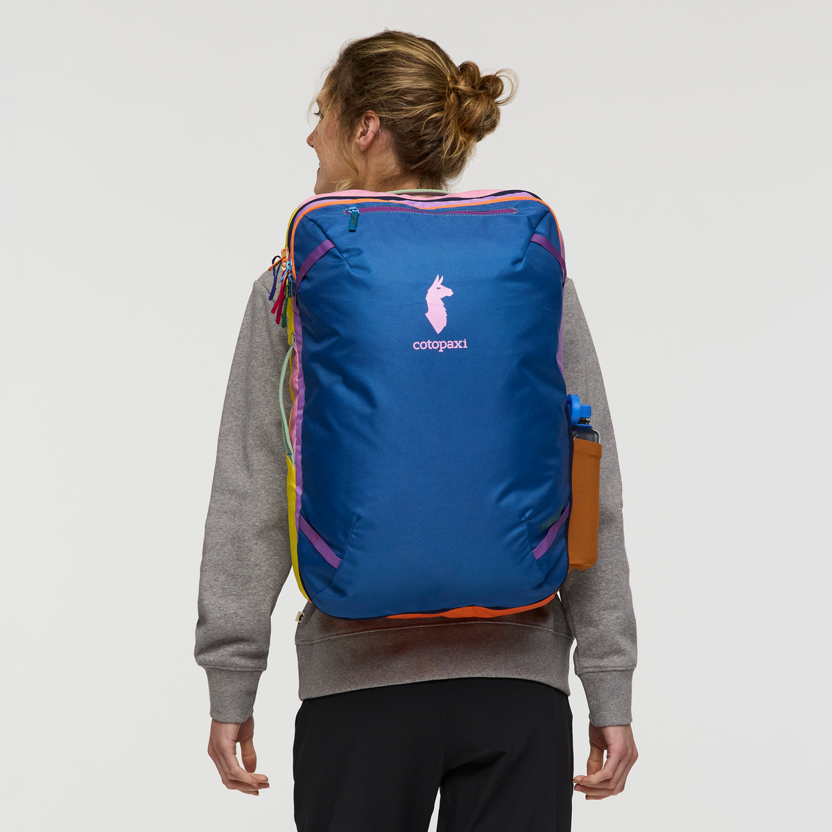 Blue travel backpack with rainbow striped accents worn by person, rear view showing logo