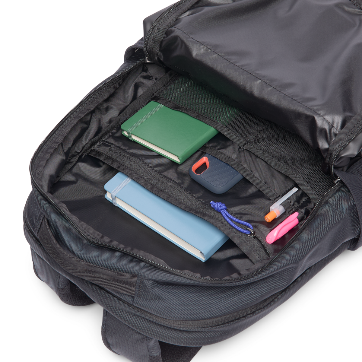 Black daypack with main compartment open showing books, external hard drive, and pens inside