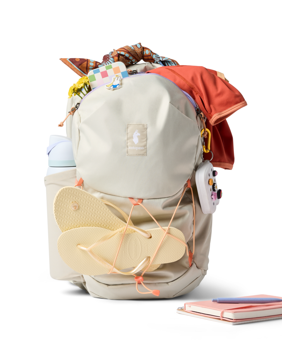 Light gray daypack with orange accents, flip-flops attached, filled with colorful items on white background