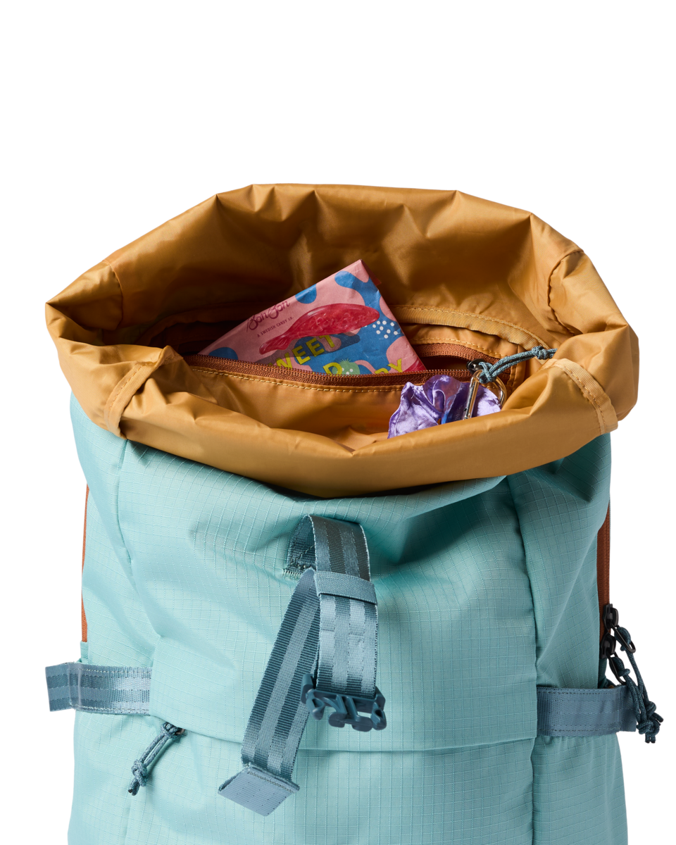 Open roll-top daypack in teal with mustard lining, showing interior zipper pocket with snacks