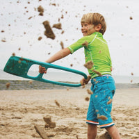 Child playing with teal colored sand scoop toy on beach, sand flying through air