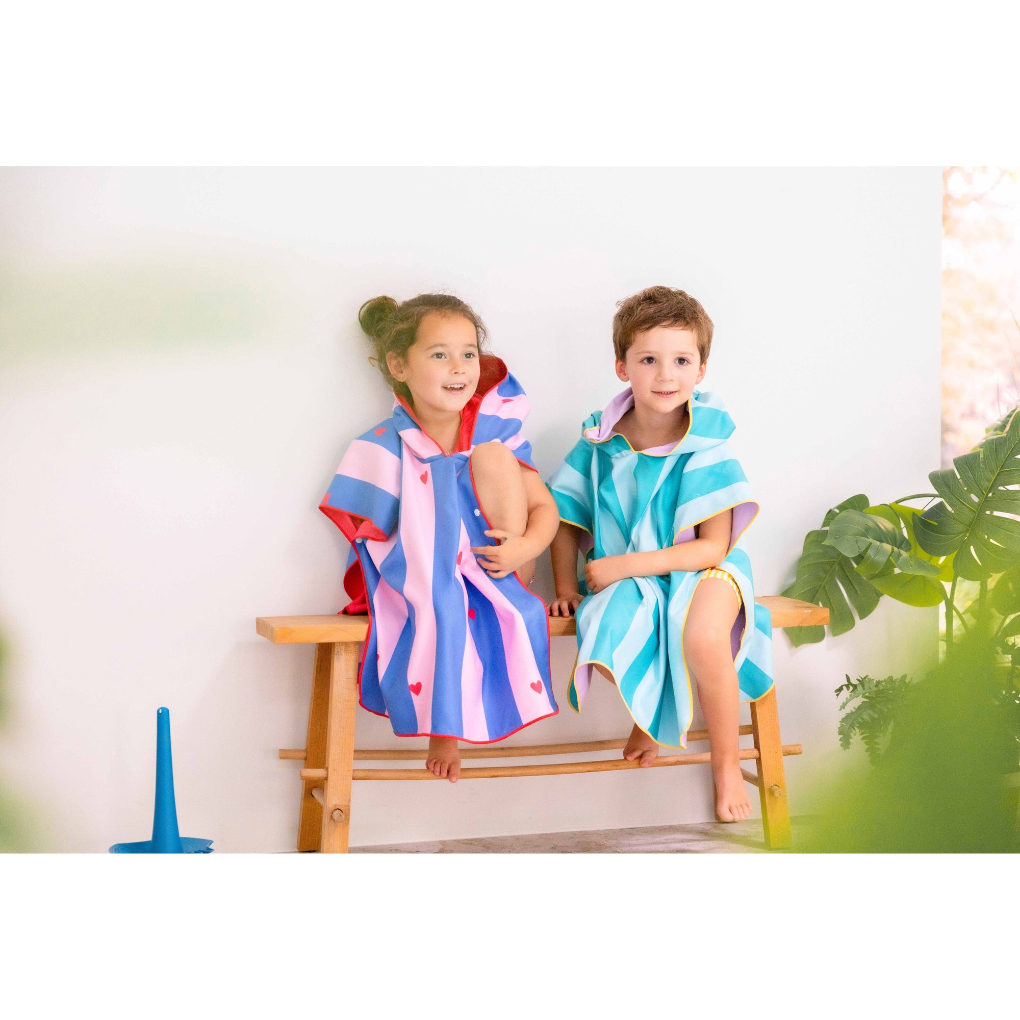 Two children wearing colorful striped hooded beach ponchos sitting on wooden bench