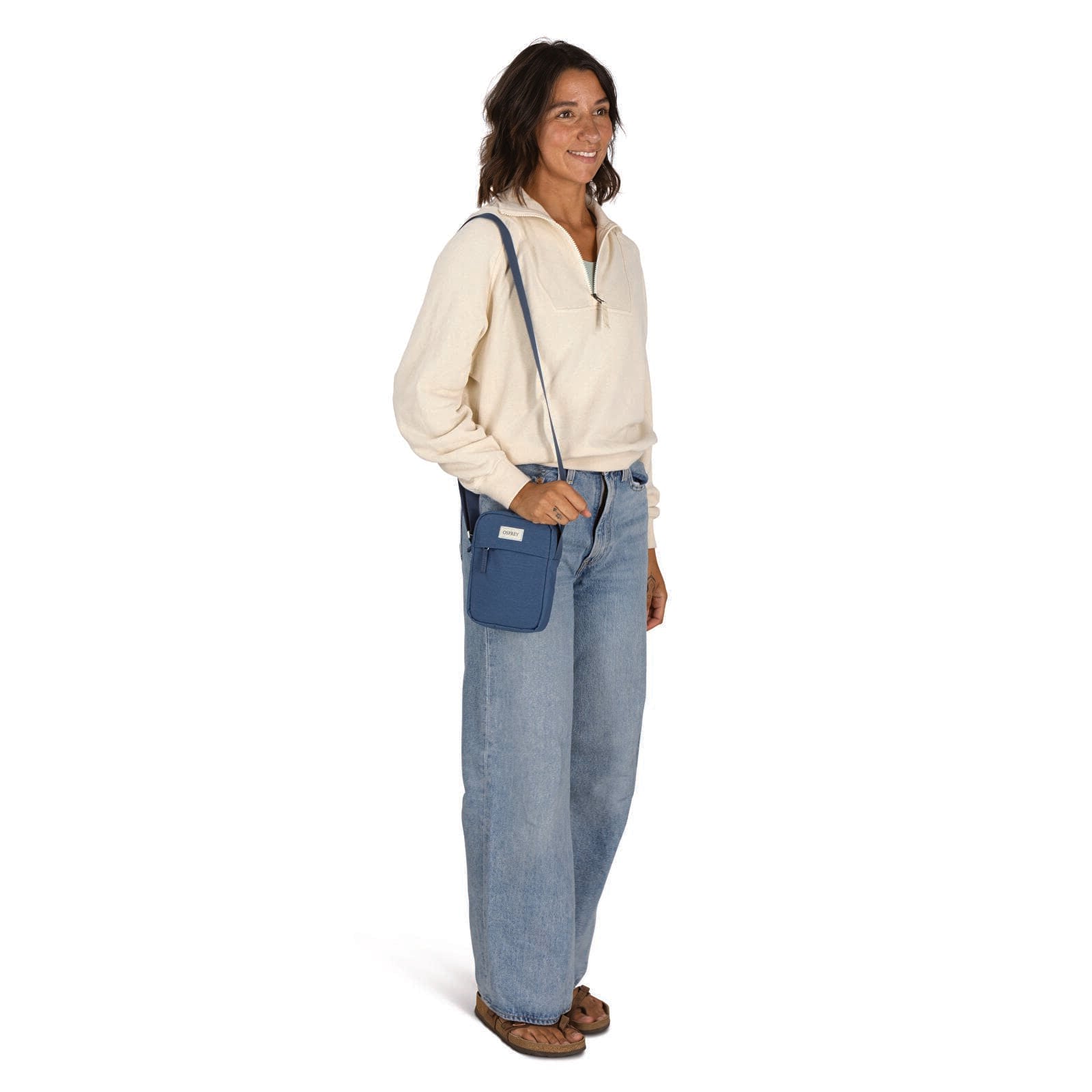 Small blue crossbody bag worn over shoulder on model in cream pullover and wide-leg jeans
