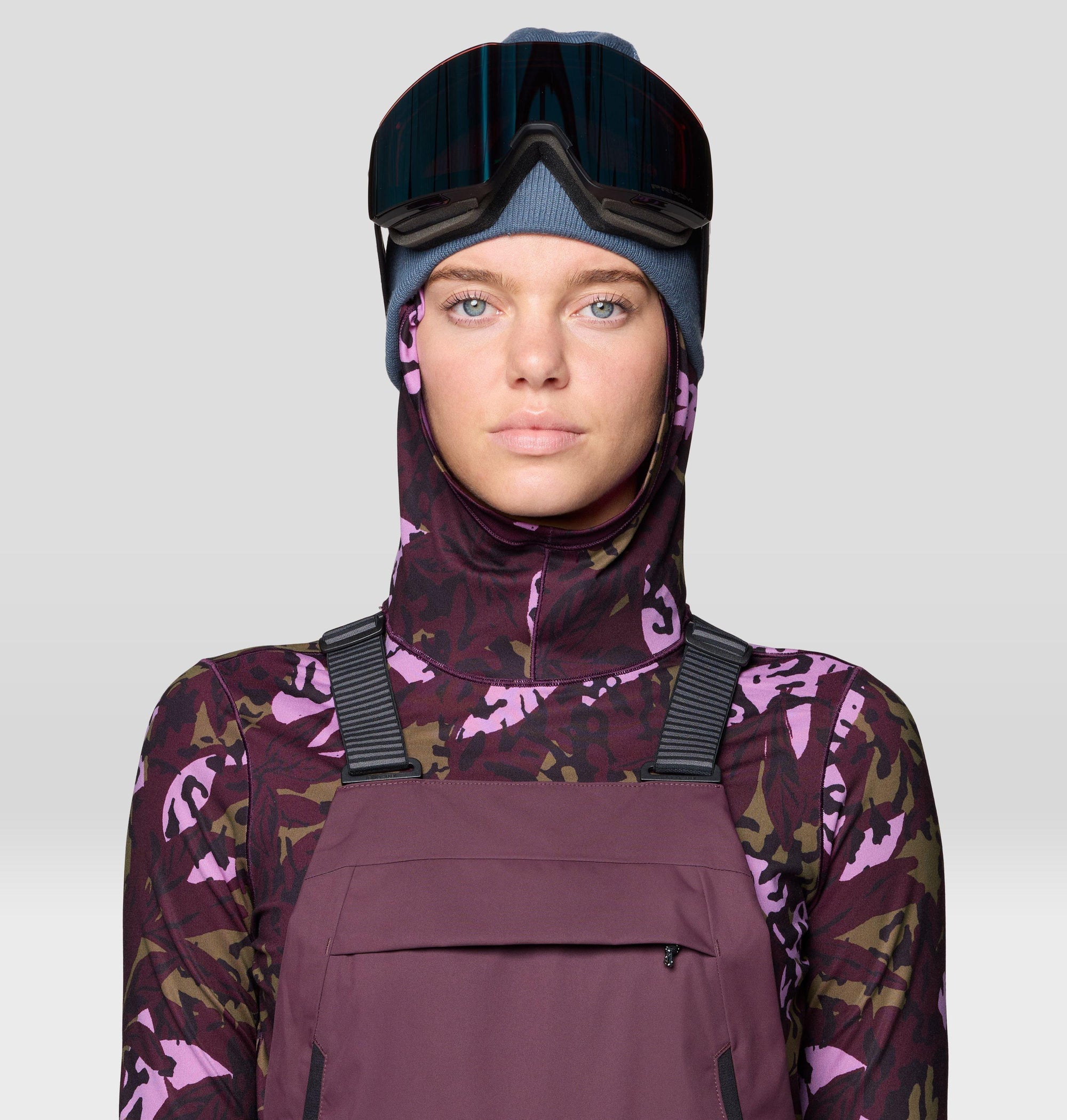 Women's ski bib overalls in plum purple with shoulder straps, worn with camo base layer and goggles