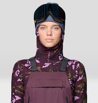 Women's ski bib overalls in plum purple with shoulder straps, worn with camo base layer and goggles