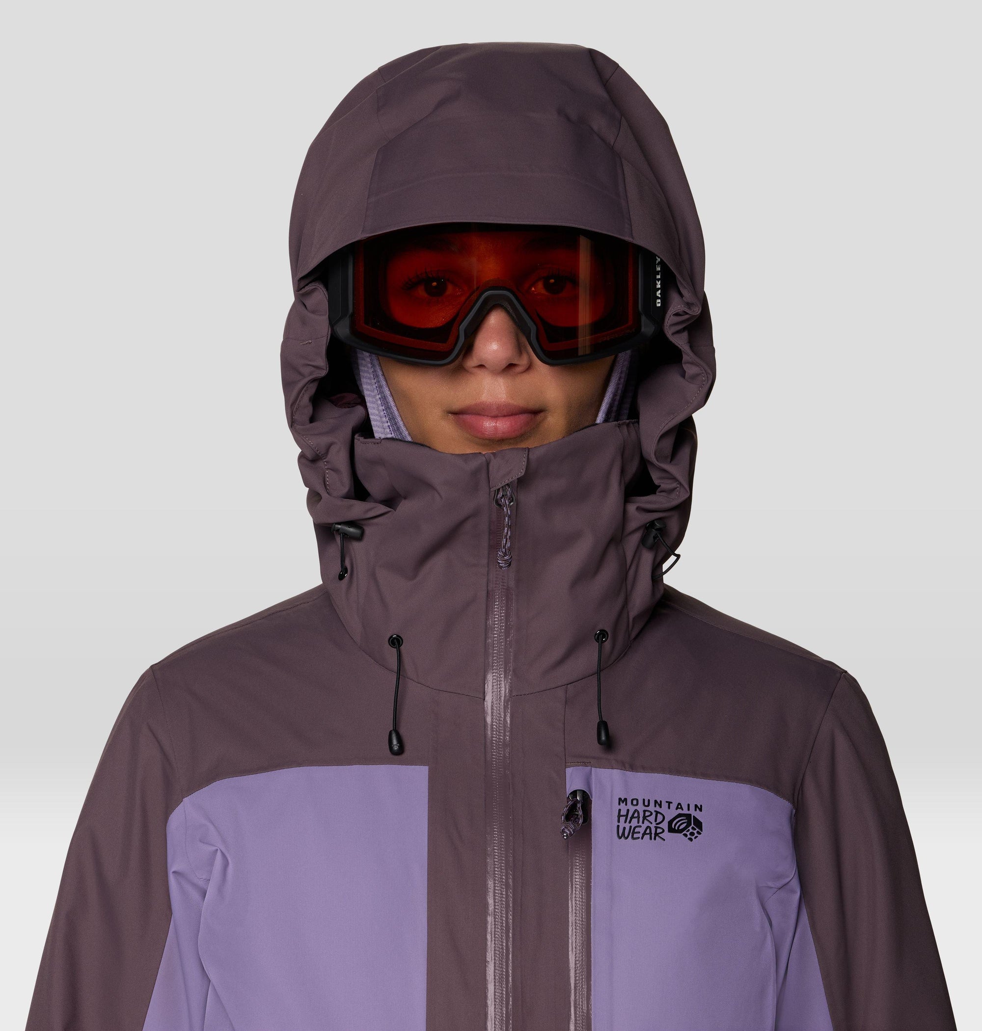 Women's ski jacket with helmet-compatible hood, adjustable collar, and two-tone purple colorblock design, close-up front view