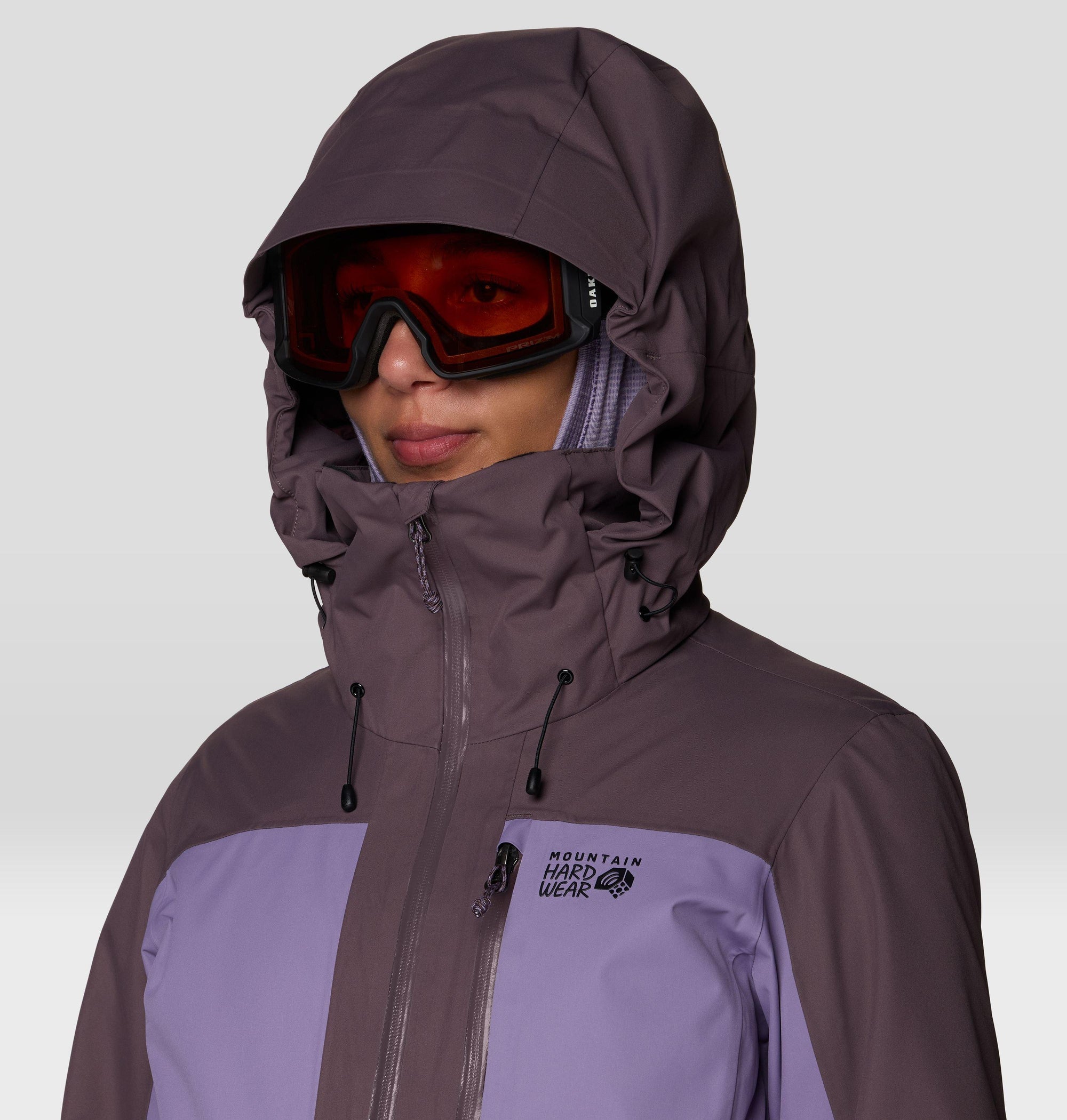 Women's hooded ski jacket with two-tone purple colorblocking, worn with orange-lens ski goggles, close-up view