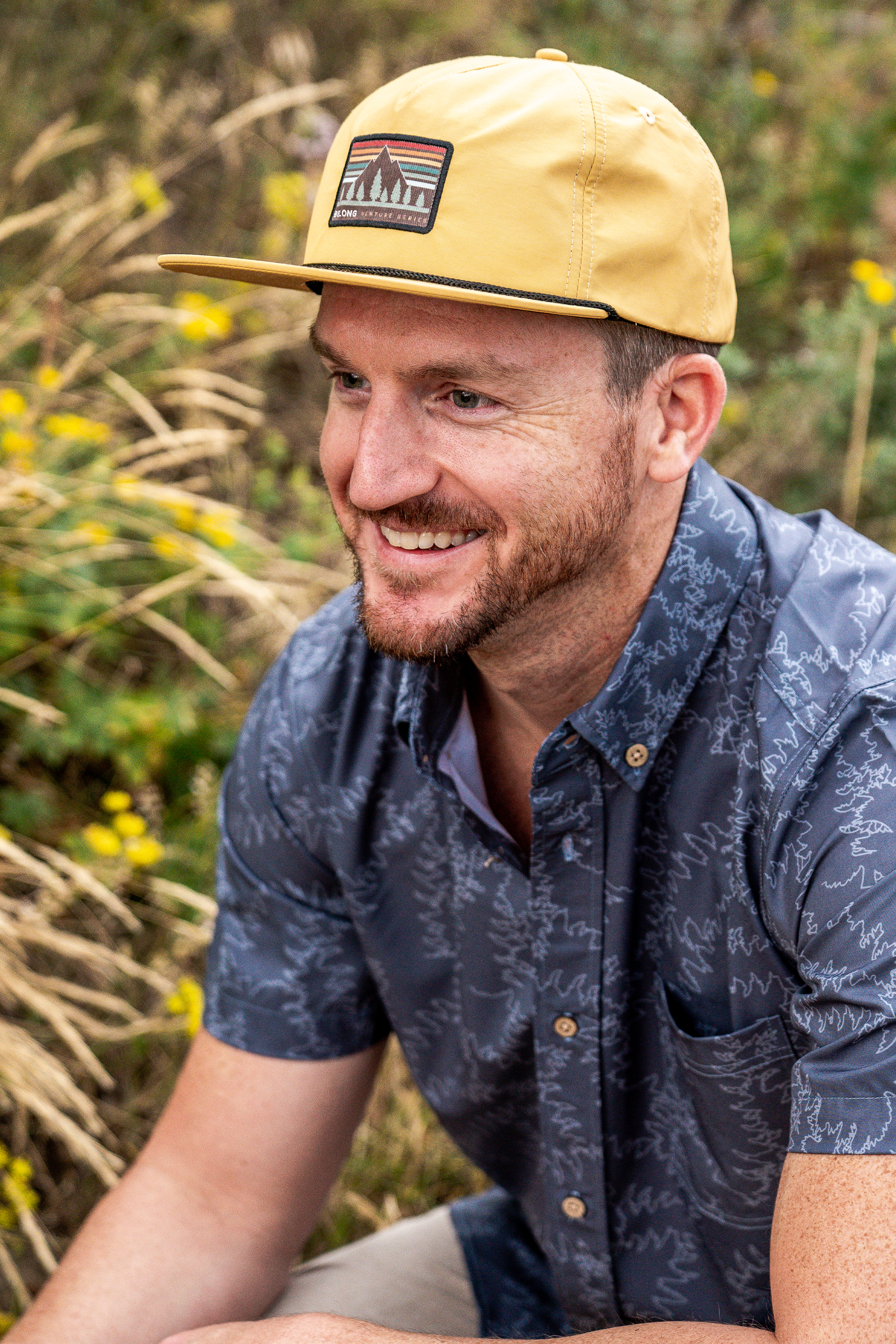Man wearing a mustard yellow snapback hat with mountain patch logo, outdoor lifestyle shot