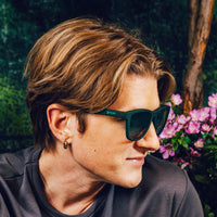 Dark green sunglasses worn by person in profile view with pink flowers in background