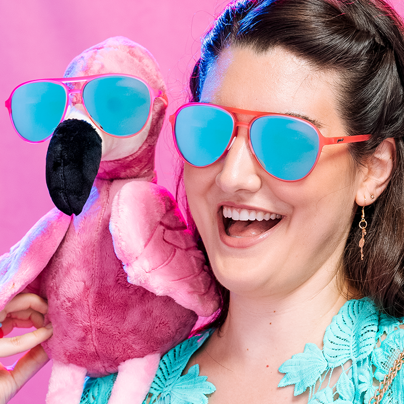 Pink aviator sunglasses with blue mirrored lenses worn by smiling woman holding plush flamingo