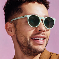 Light green round sunglasses with dark lenses worn by smiling man against pink background