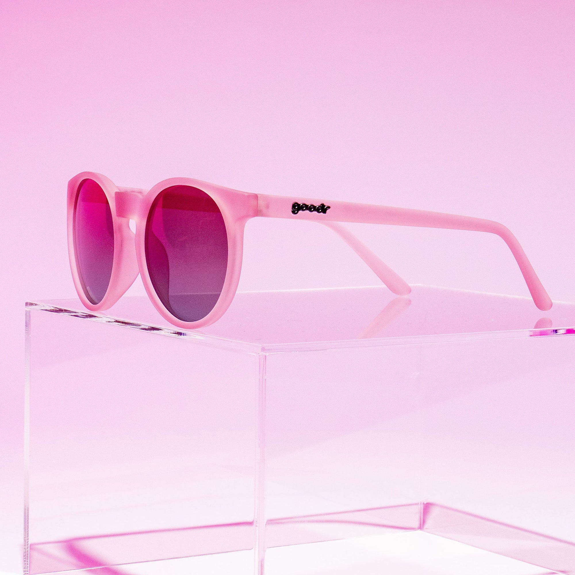 Pink round sunglasses with dark purple lenses displayed on clear acrylic stand