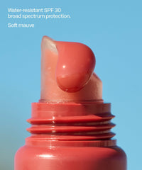 Coral-red tinted lip balm stick with SPF 30, shown extended from tube against blue background