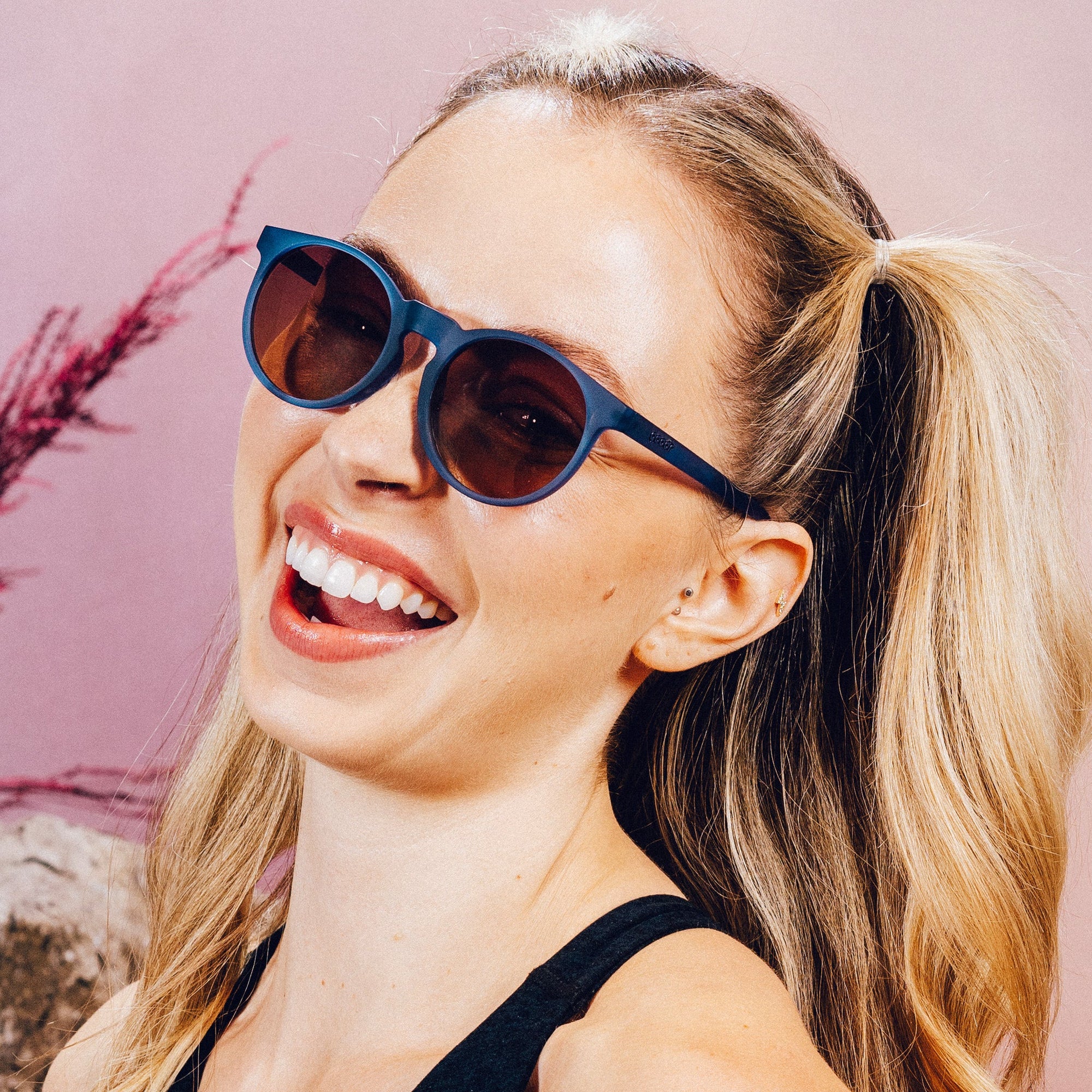 Round blue sunglasses with dark tinted lenses worn by smiling woman with blonde ponytail