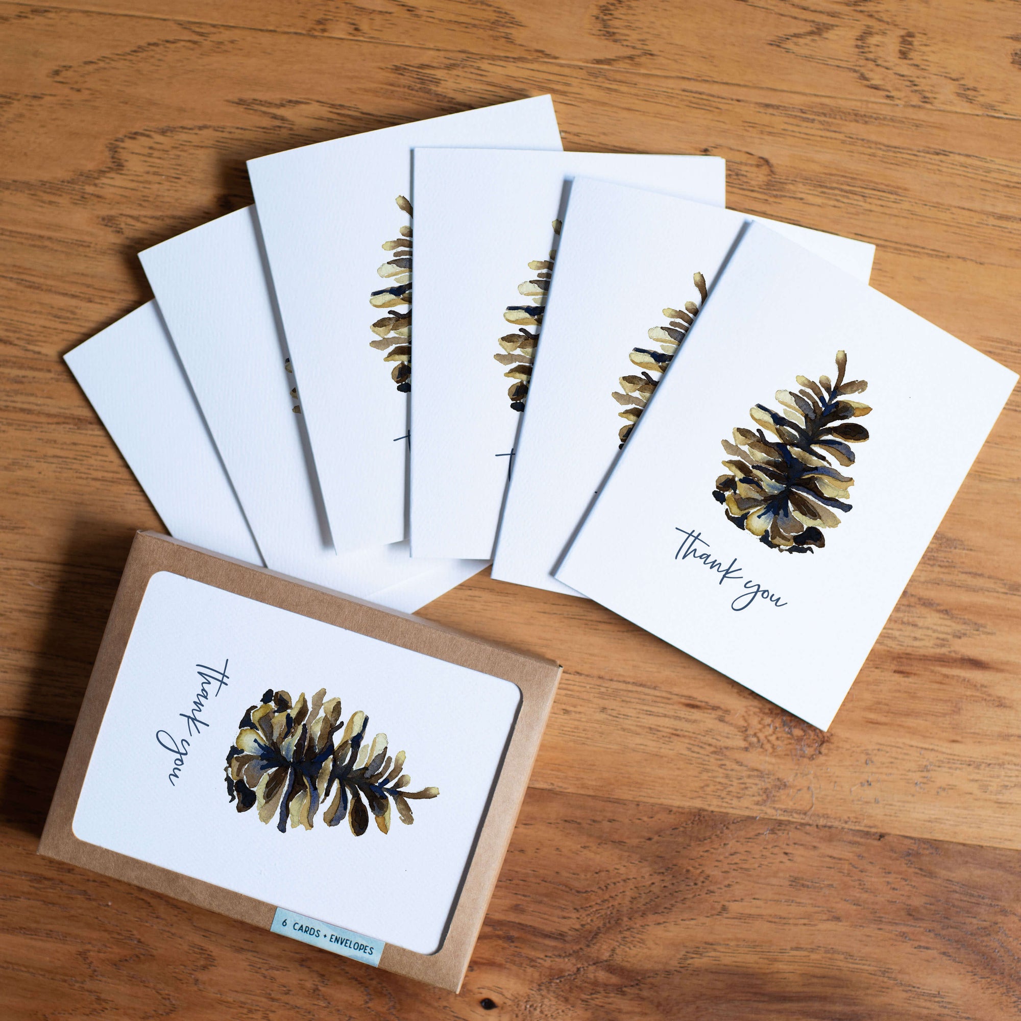 Set of thank you greeting cards with watercolor pinecone design displayed with brown packaging box