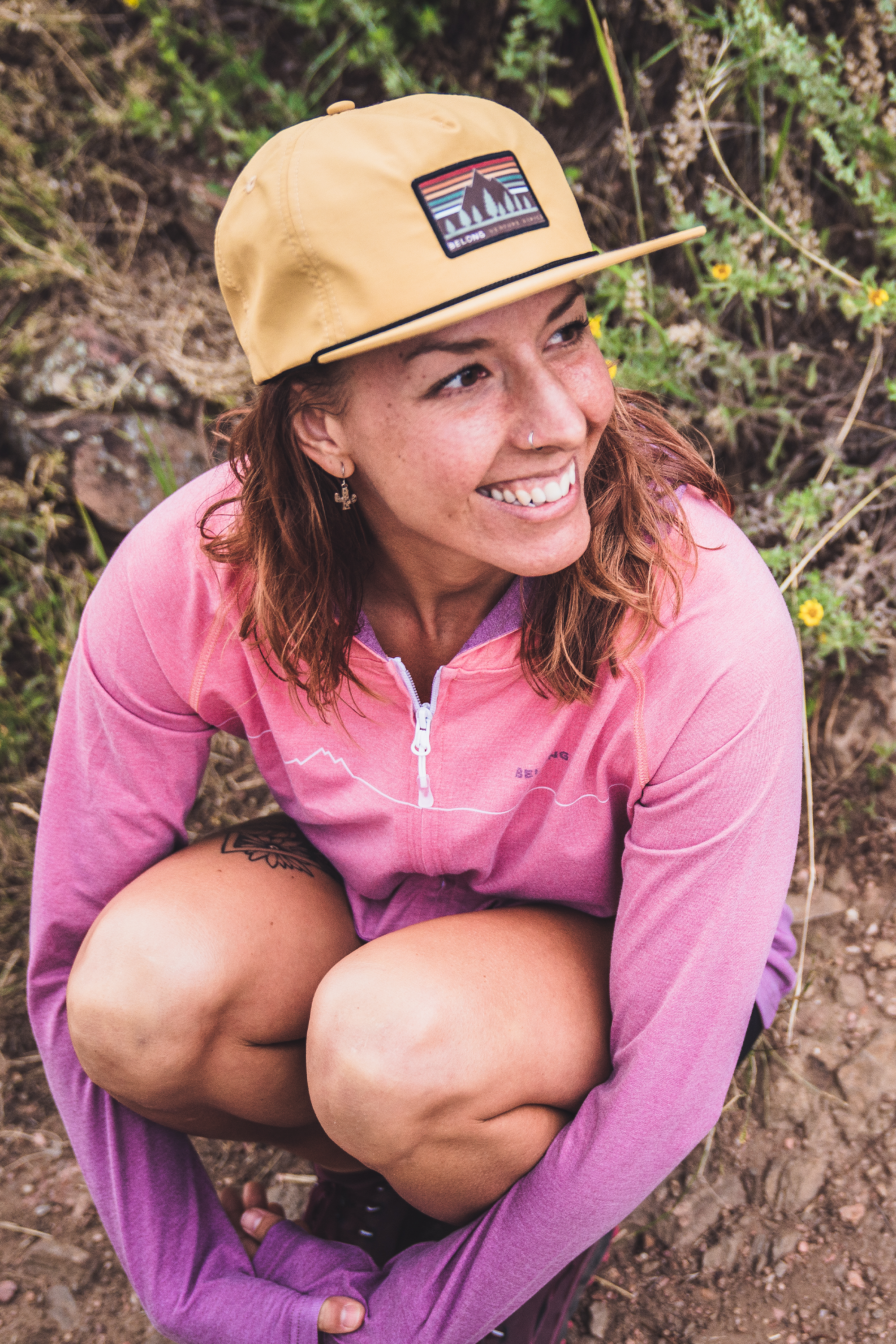 Woman wearing a mustard yellow flat-brim snapback hat with mountain logo patch, smiling outdoors
