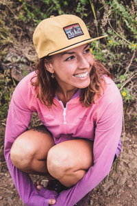 Woman wearing a mustard yellow flat-brim snapback hat with mountain logo patch, smiling outdoors