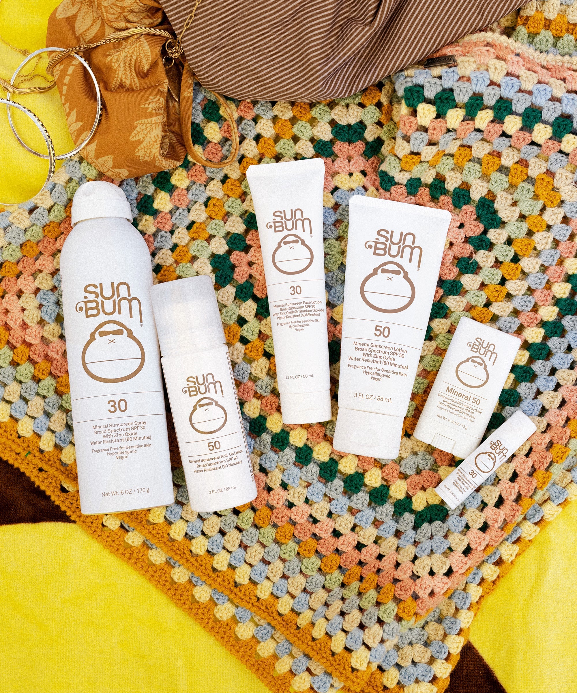 Collection of white sunscreen products including spray bottles and lotion tubes arranged on colorful crocheted blanket