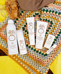Collection of white sunscreen products including spray bottles and lotion tubes arranged on colorful crocheted blanket