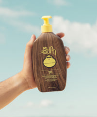 Hand holding brown wood-textured sunscreen lotion bottle with yellow pump dispenser against sky background