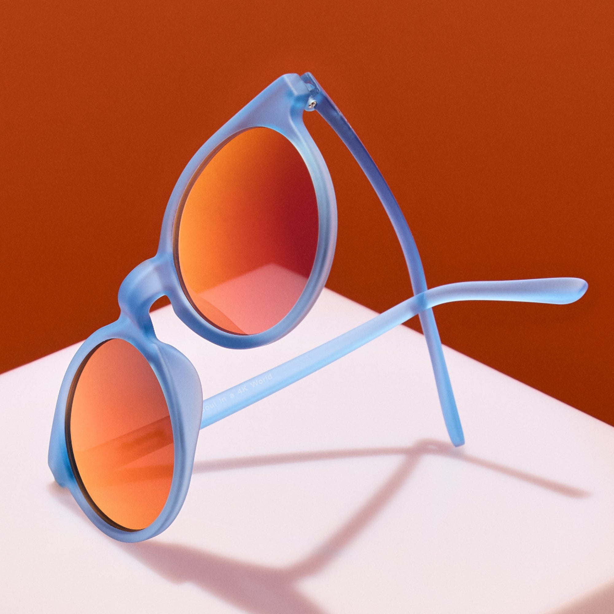 Translucent blue sunglasses with orange gradient lenses displayed on white platform against orange background