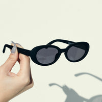 Black oval sunglasses with dark lenses held in hand against neutral background