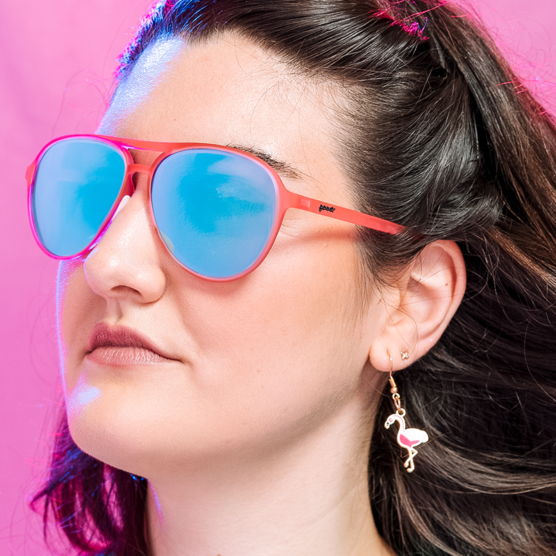 Pink aviator sunglasses with blue mirrored lenses worn by woman against pink background