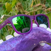 Purple sunglasses with green mirrored lenses resting on purple crystal with foliage background