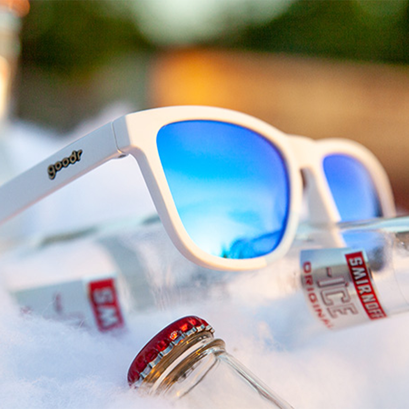 White framed sunglasses with blue mirrored lenses displayed on ice with beverage cans
