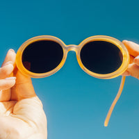 Round translucent yellow sunglasses with dark lenses held up against blue sky background