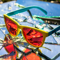 Yellow-framed sunglasses with red mirrored lenses sitting on reflective broken mirror surface