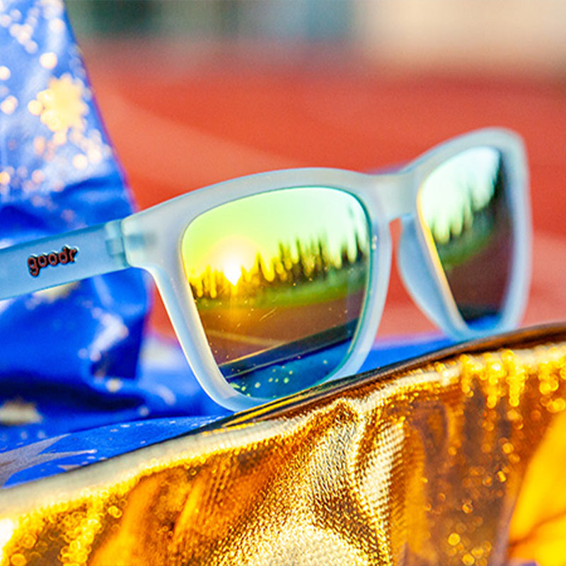Clear blue framed sunglasses with yellow-green mirrored lenses resting on golden metallic fabric