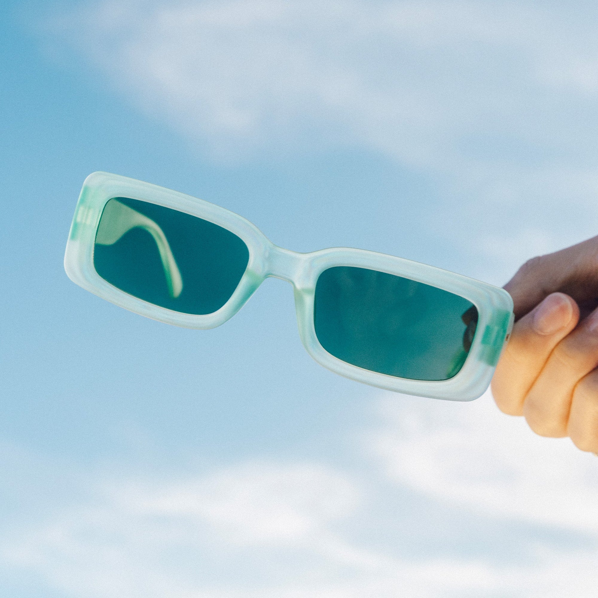 Translucent mint green rectangular sunglasses with dark teal lenses held against blue sky