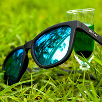 Black sunglasses with blue-green mirrored lenses resting on grass beside clear glass