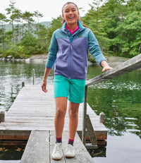Girl wearing colorblock fleece jacket with purple body and teal sleeves, standing on wooden dock