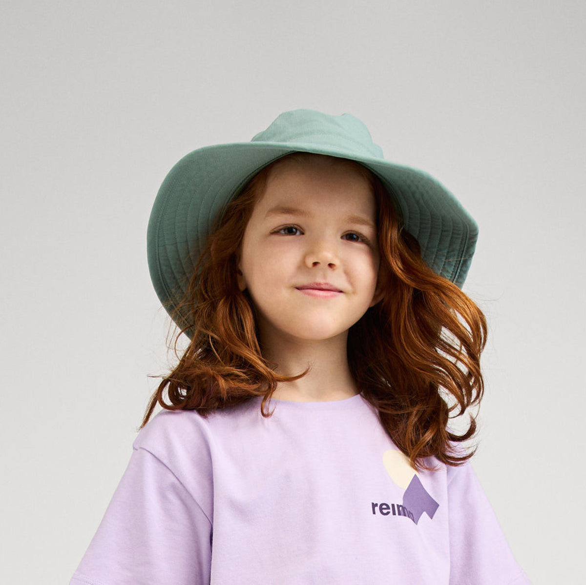 Child wearing sage green wide-brimmed sun hat with wavy hair in studio portrait