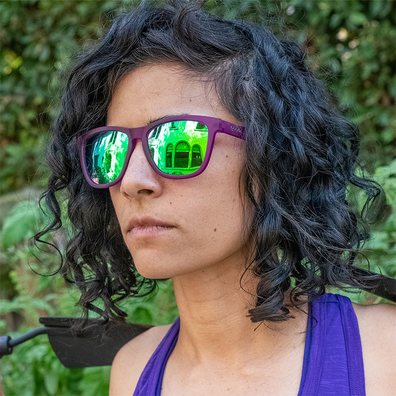 Purple-framed sunglasses with green mirrored lenses worn by woman with curly dark hair