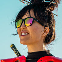 Black framed sunglasses with rainbow reflective lenses worn by smiling woman outdoors