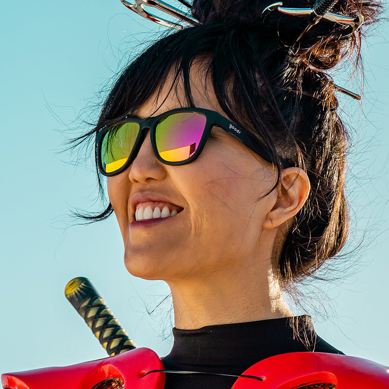 Black framed sunglasses with rainbow reflective lenses worn by smiling woman outdoors