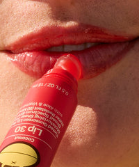 Coral-red tinted lip balm stick being applied to lips in close-up lifestyle shot