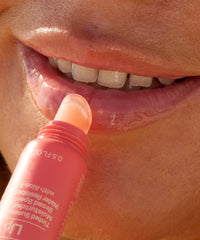 Coral pink tinted lip balm stick being applied to lips in close-up lifestyle shot