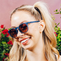Round blue sunglasses with brown lenses worn by smiling woman with blonde ponytail