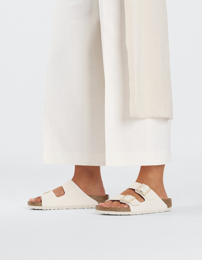 Cream colored two-strap sandals with buckles and cork footbed, worn with white pants